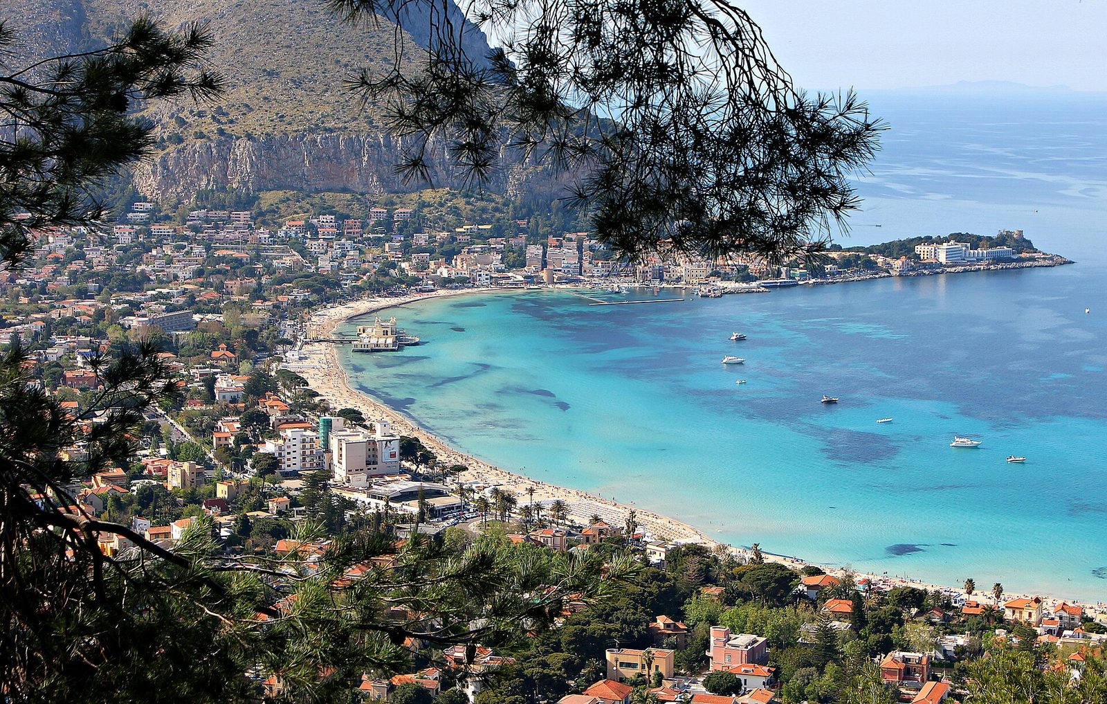Il golfo di Mondello visto dall'alto tra i rami di pino: la spiaggia a mezzaluna, l'acqua turchese, l'edificio liberty sul mare e la montagna alle spalle
